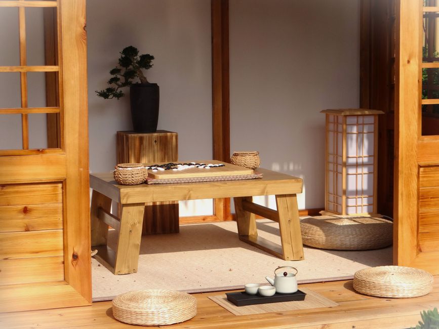 Tranquil Japanese Zen room featuring a Go board, tea set, bonsai tree, and wicker cushions on a wooden table.
